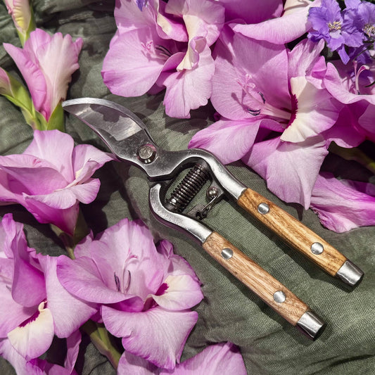 A pair of pruning shears with wooden handles and silver metal blades, arranged on a dark textured fabric surrounded by pink and purple flowers.