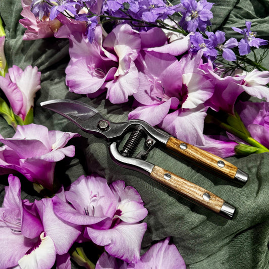 A pair of hand pruners with wooden handles and stainless steel blades, styled on a dark green fabric background surrounded by purple flowers.