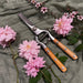 A pair of pruning shears with light brown wooden handles and silver metal blades, arranged on a textured olive green fabric with pink roses and green leaves.