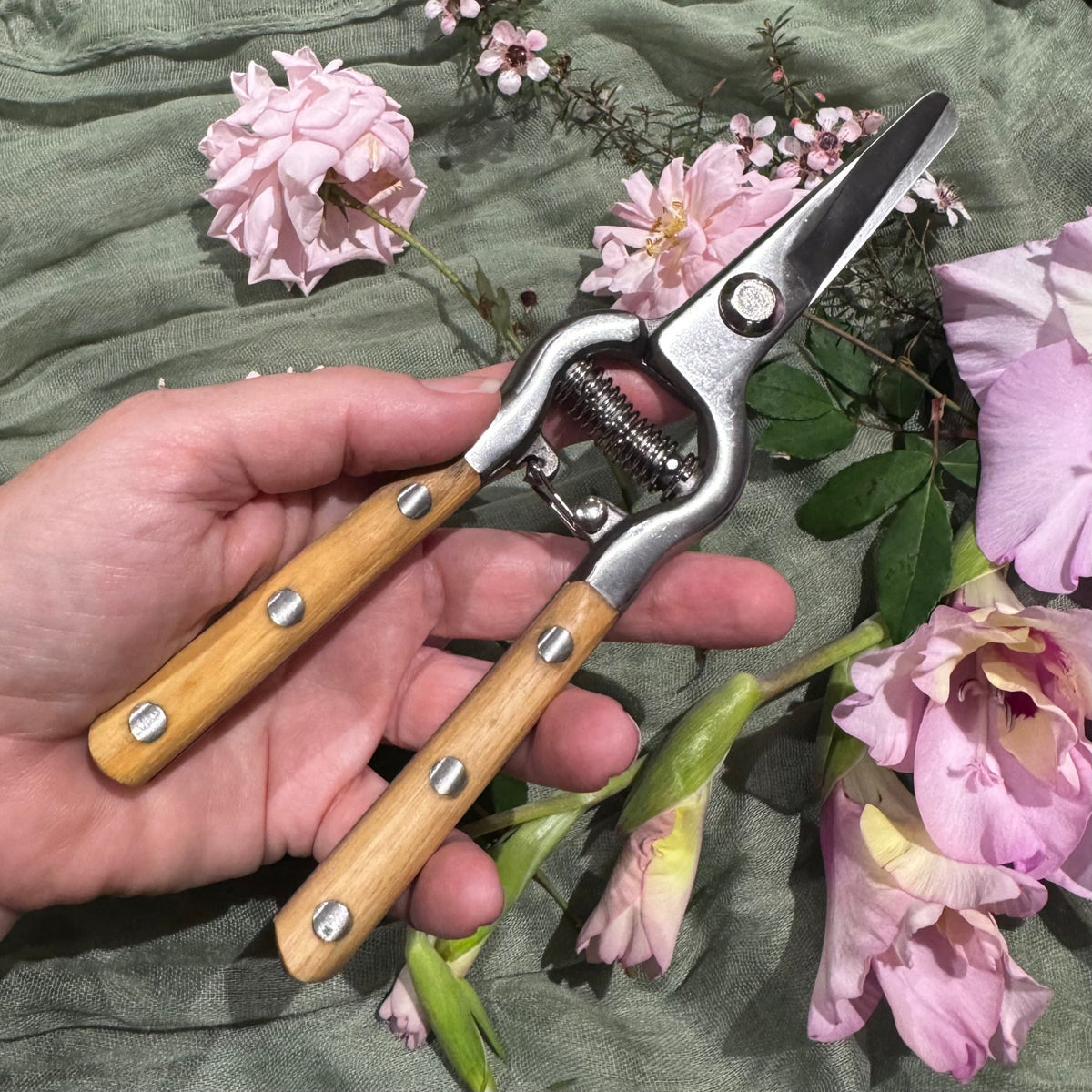 A hand holding a pair of pruning shears with light wood handles and silver metal blades, surrounded by pink roses on a textured sage green fabric background.